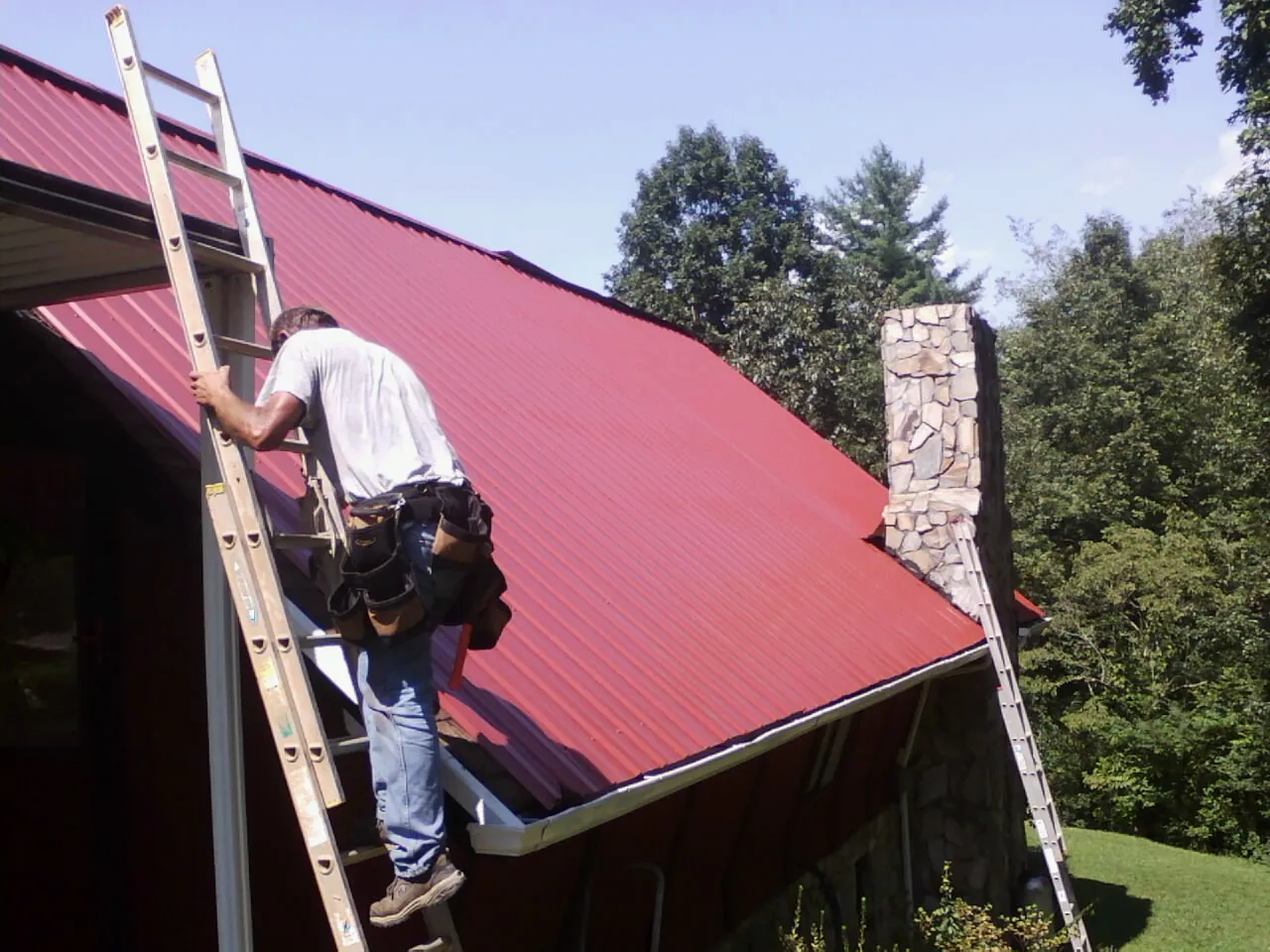 Roofer on a ladder beside a red metal roof and stone chimney — Soffit & Fascia Repair by Roofing Brothers in Ponderay